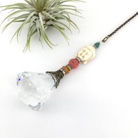 This charming handmade crystal hanging will adorn any room with rainbows and lively reflections, making it a delightful addition for yourself or a thoughtful gift that will be treasured.Featuring a high-quality, medium (40mm) faceted crystal prism, meticulously hand-strung with Howlite gemstone Buddha Focal, Cherry Quartz and distinctive Czech glass beads. It is completed with chain and hook, ensuring effortless hanging and allowing gentle movement that makes the rainbows dance.