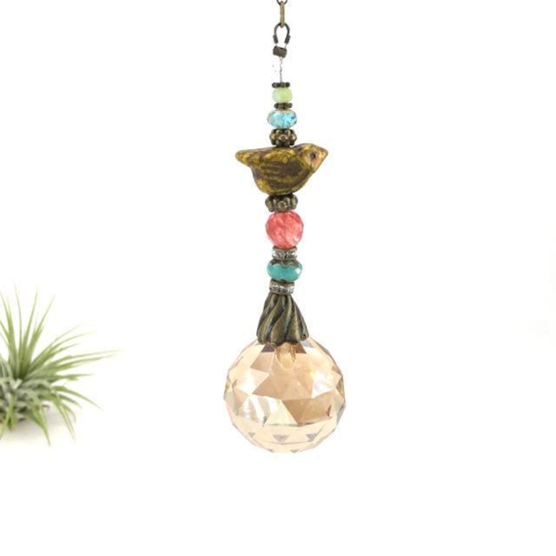 This charming handmade crystal sun catcher hanging will adorn any room with rainbows and bring lively reflections. Simple and elegant, our unique design features a high-quality Asfour Honey AB (30mm) faceted crystal prism sphere, meticulously hand-strung with unique beads. Czech Glass bird focal, vintage Czech Glass beads and Cherry Quartz gemstone. It is completed with chain and hook, ensuring effortless hanging and allowing gentle movement that makes the rainbows dance. Hanging Length of 17".