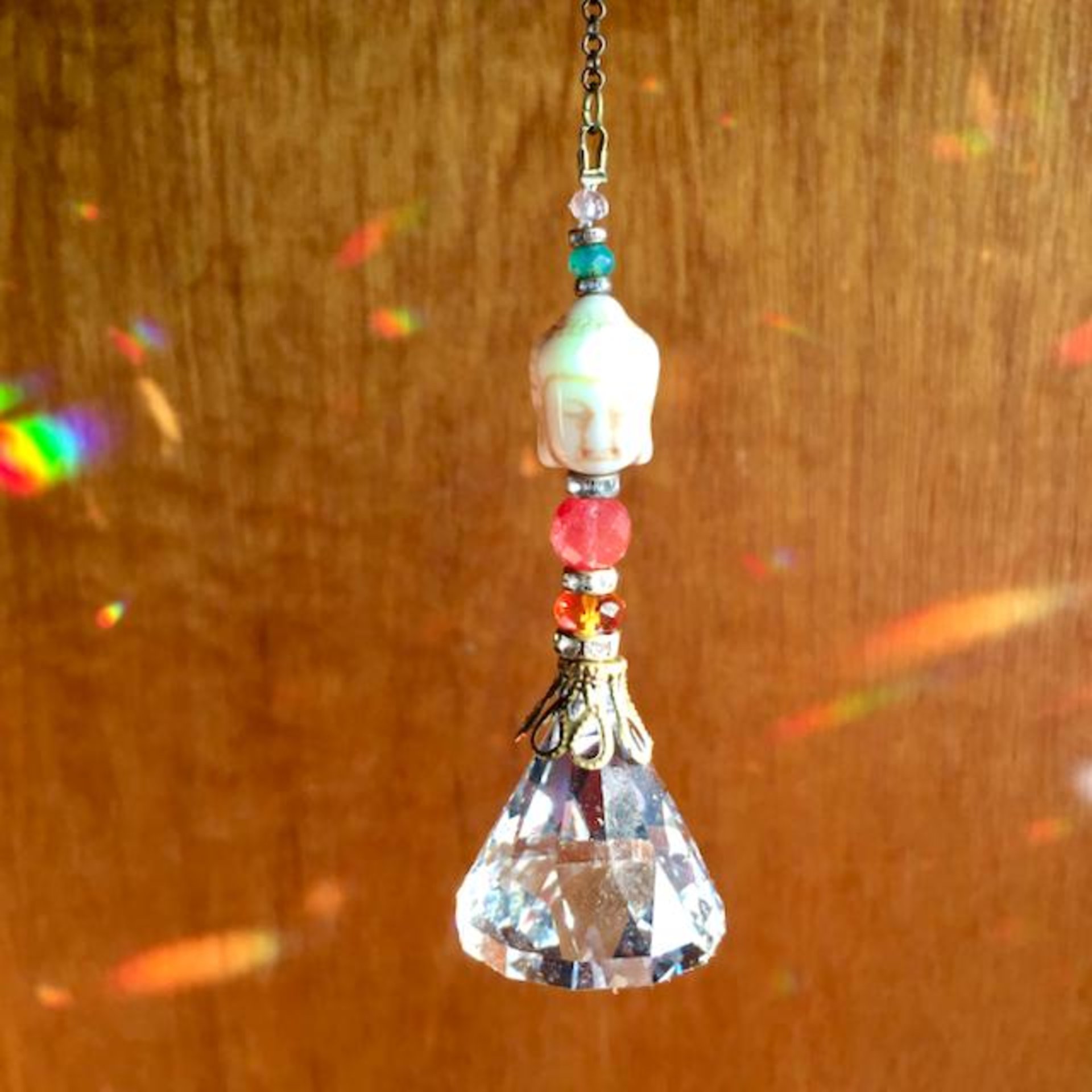 This charming handmade crystal hanging will adorn any room with rainbows and lively reflections, making it a delightful addition for yourself or a thoughtful gift that will be treasured.Featuring a high-quality, medium (40mm) faceted crystal prism, meticulously hand-strung with Howlite gemstone Buddha Focal, Cherry Quartz and distinctive Czech glass beads. It is completed with chain and hook, ensuring effortless hanging and allowing gentle movement that makes the rainbows dance.