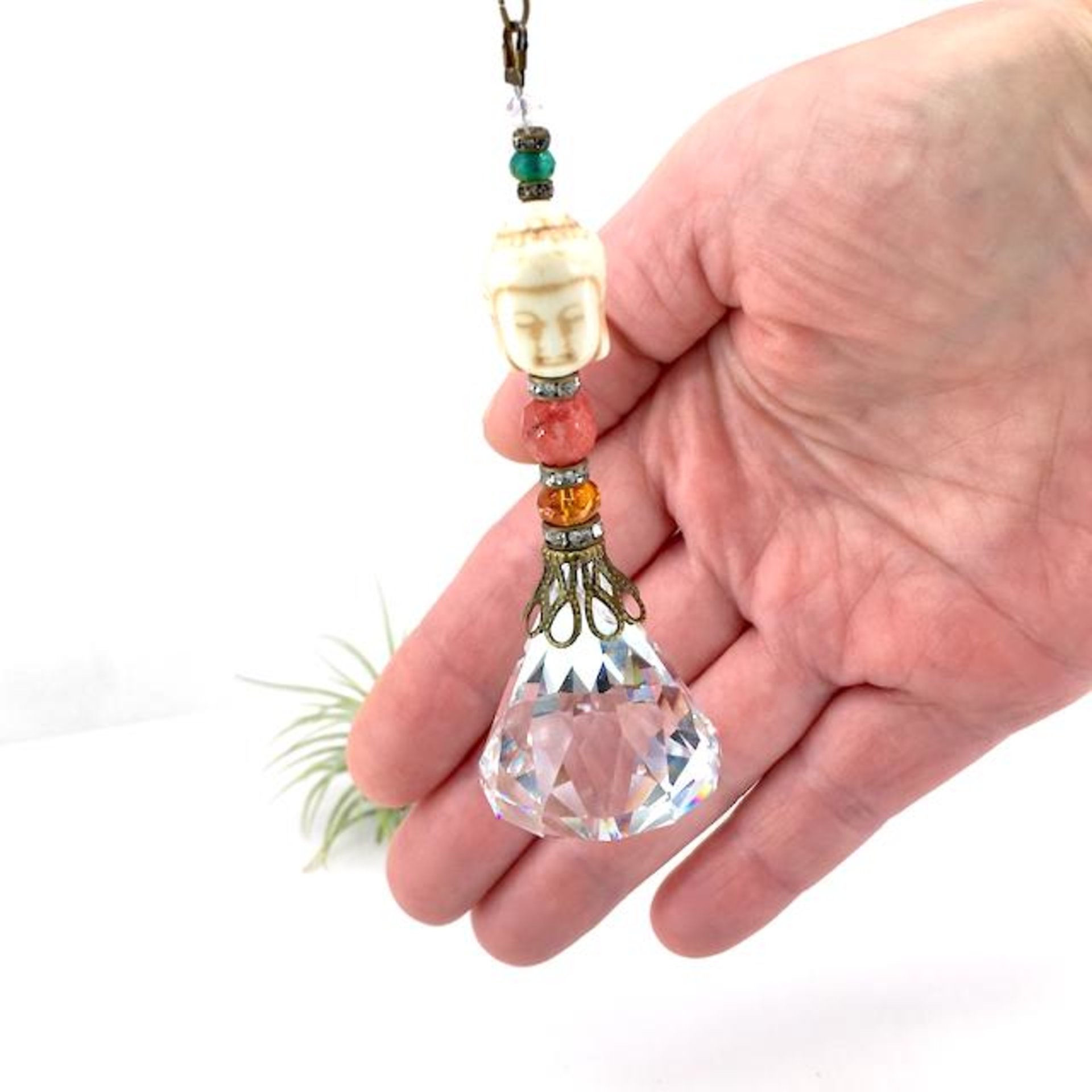 This charming handmade crystal hanging will adorn any room with rainbows and lively reflections, making it a delightful addition for yourself or a thoughtful gift that will be treasured.Featuring a high-quality, medium (40mm) faceted crystal prism, meticulously hand-strung with Howlite gemstone Buddha Focal, Cherry Quartz and distinctive Czech glass beads. It is completed with chain and hook, ensuring effortless hanging and allowing gentle movement that makes the rainbows dance.